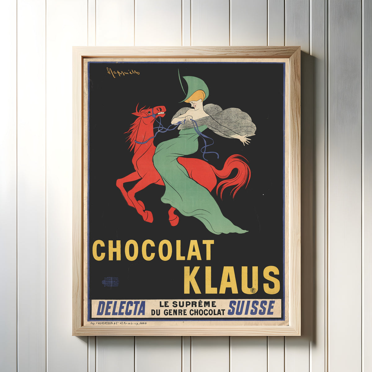 Framed art print of Chocolat Klaus by Leonetto Cappiello, in a light wood frame on a white panelled wall