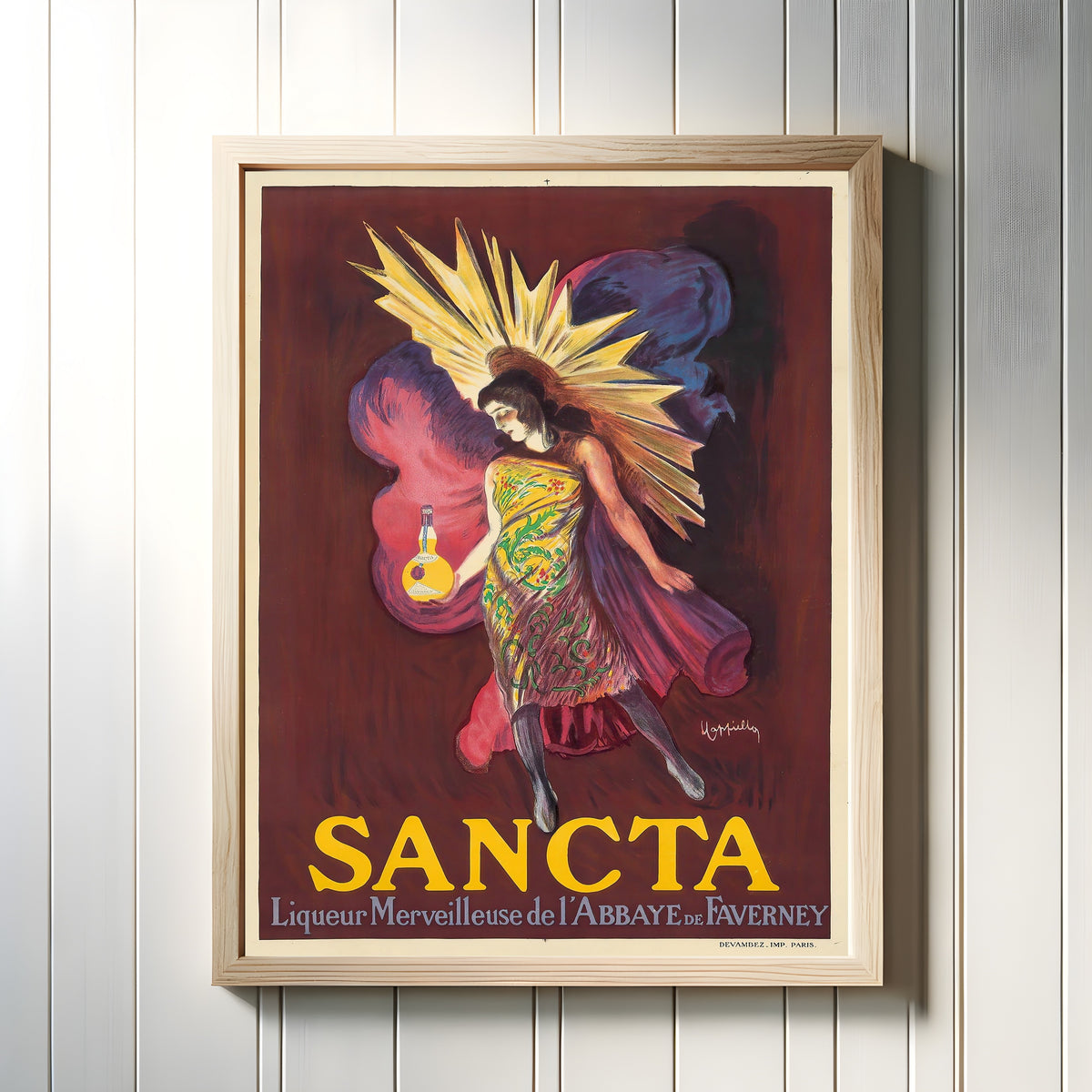 Framed art print of Sancta Liquor by Leonetto Cappiello, in a light wood frame on a white panelled wall