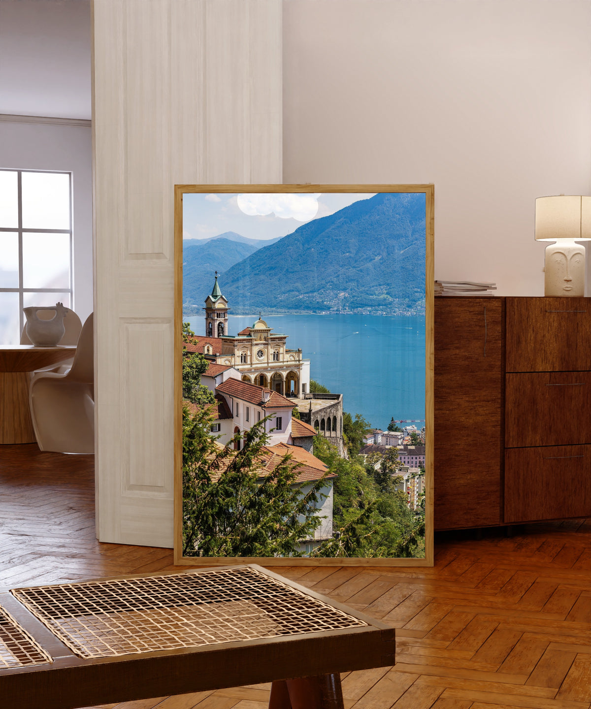 Framed art print of Madonna del Sasso in Orselina by Daniela Faust, in a light wood frame leaning in a warm room with a parquet floor