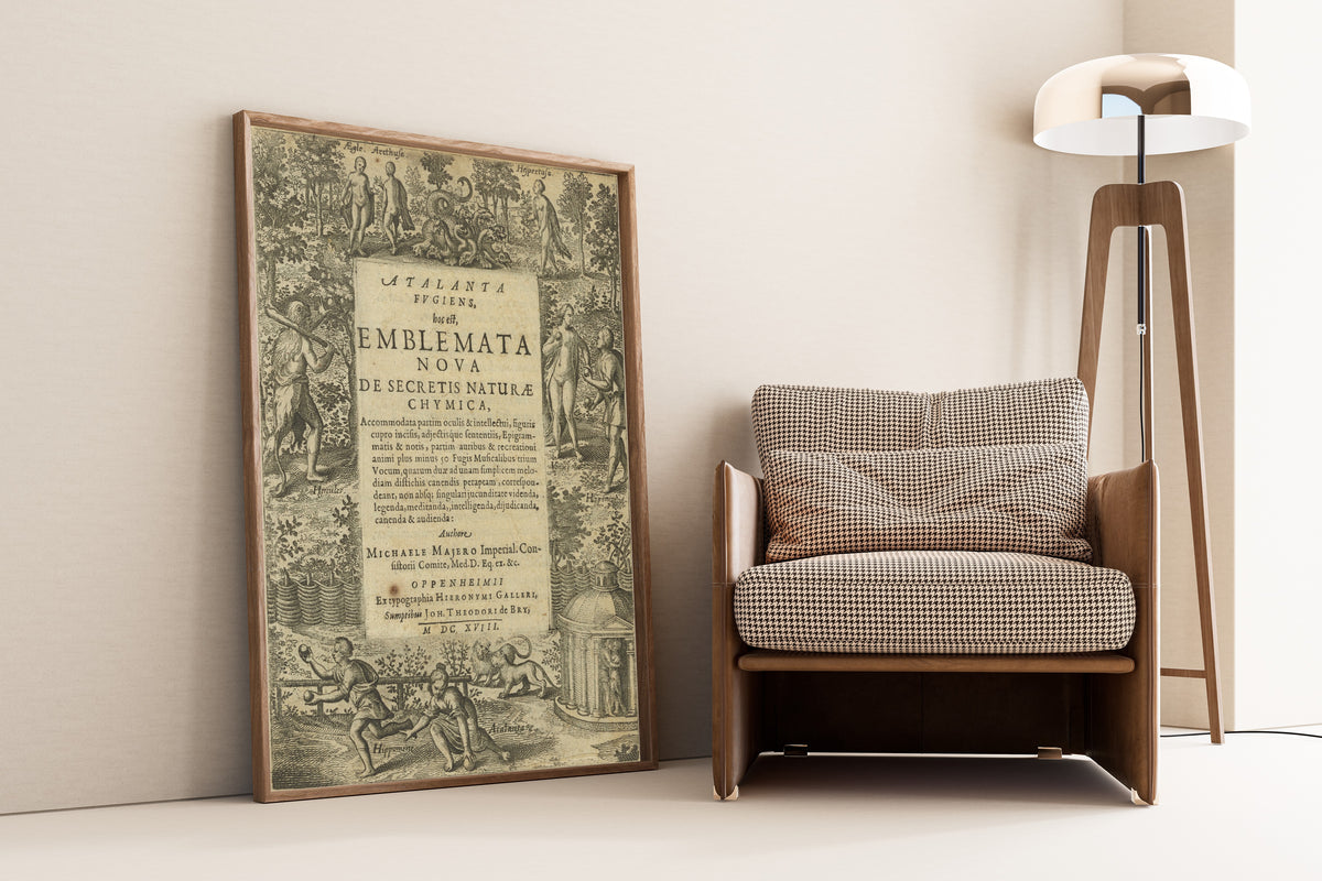 Framed art print of Atalanta Fugiens, Title Page by Michael Maier, leaning against a wall beside an armchair and a floor lamp