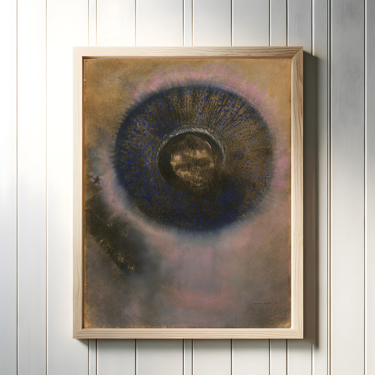 Framed art print of Head Within an Aureole (1895) by Odilon Redon, in a light wood frame on a white panelled wall