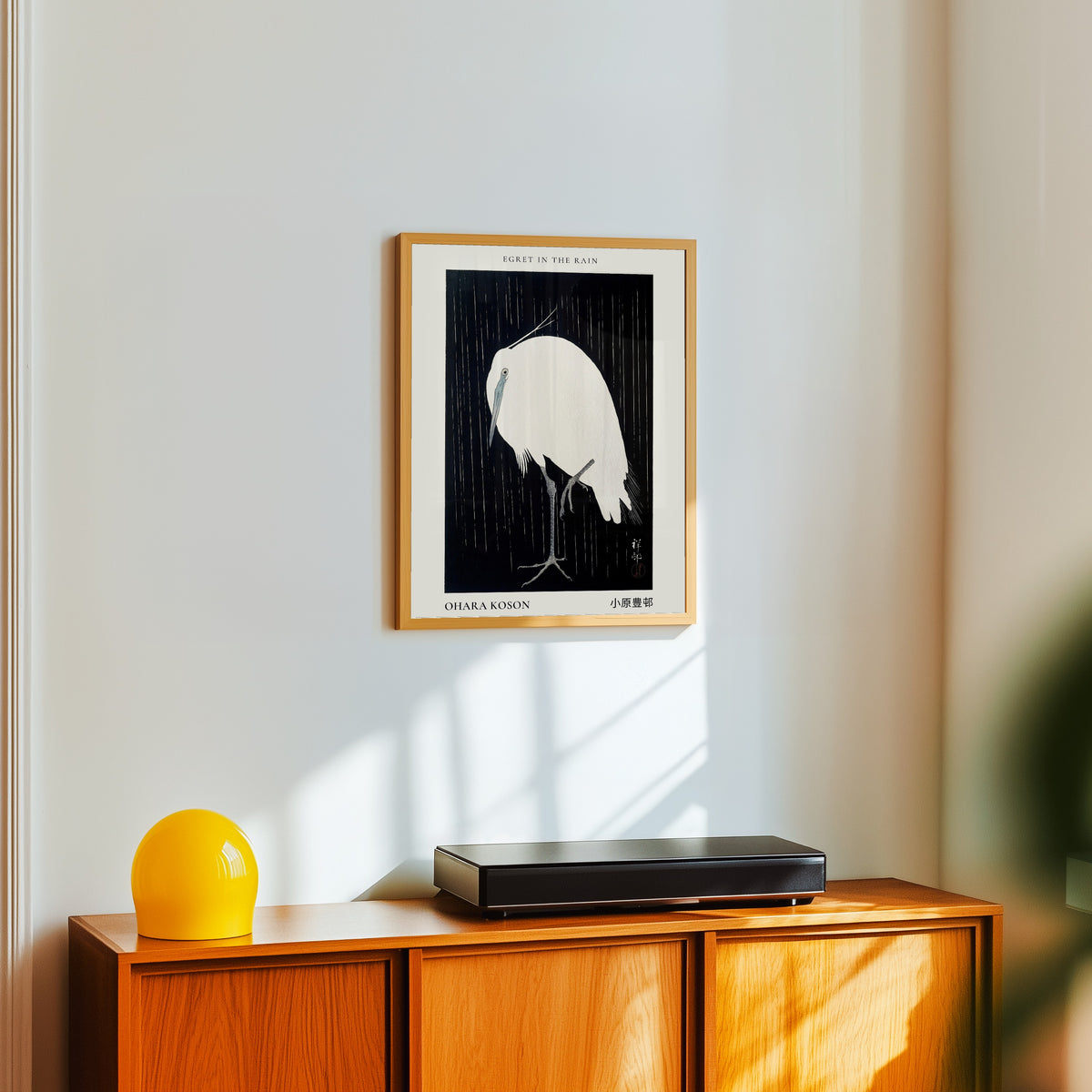 Framed art print of Egret in the Rain by Ohara Koson, in a light wood frame on a white wall above an orange sideboard