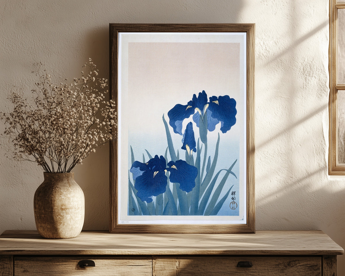 Framed art print of Irises in Blue by Ohara Koson, in a dark wood frame leaning on a rustic console beside dried flowers