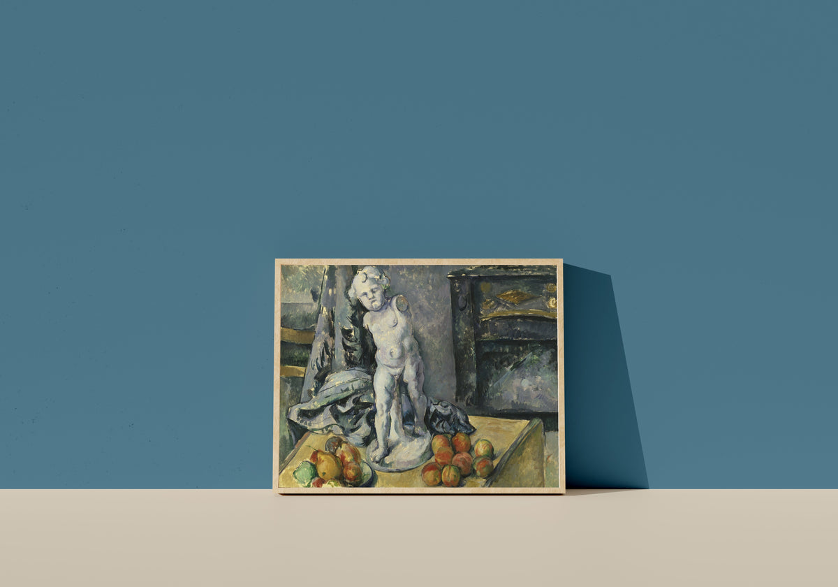 Framed art print of Still Life with Plaster Cupid by Paul Cezanne, in a light wood frame leaning on a blue wall