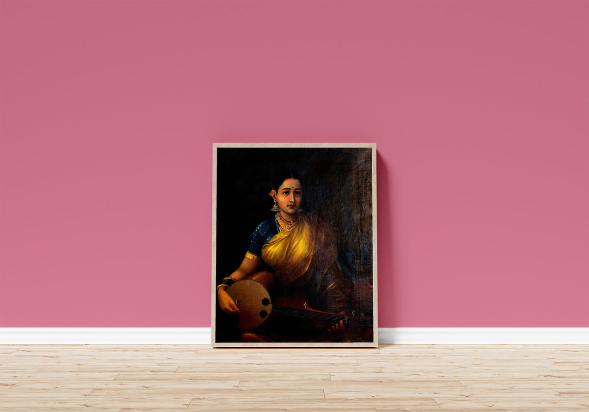 Framed art print of A Tamil Lady Playing the Swarbat by Raja Ravi Varma, in a light wood frame leaning on a pink wall above a wooden floor