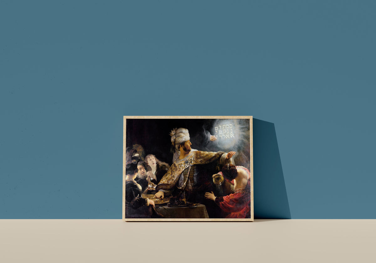 Framed art print of Belshazzar's Feast by Rembrandt van Rijn, in a light wood frame leaning on a blue wall