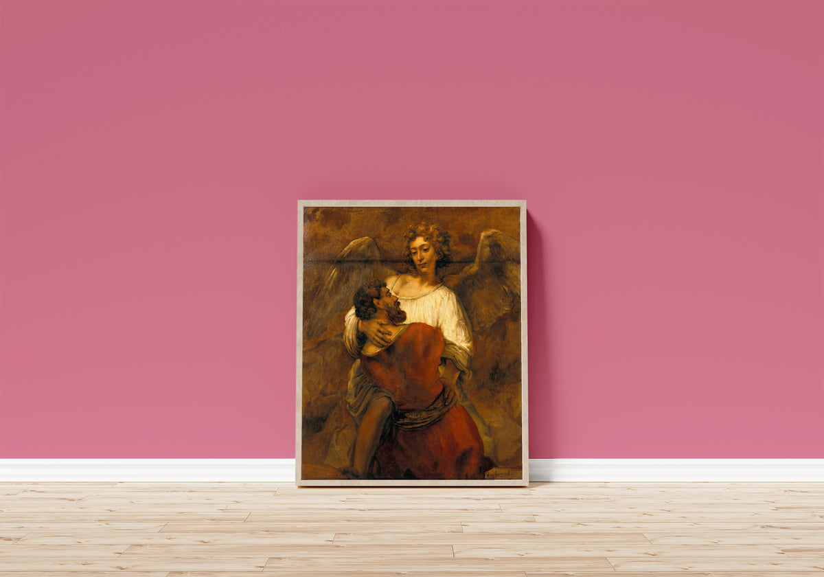 Framed art print of Jacob Wrestling with the Angel by Rembrandt van Rijn, in a light wood frame leaning on a pink wall above a wooden floor