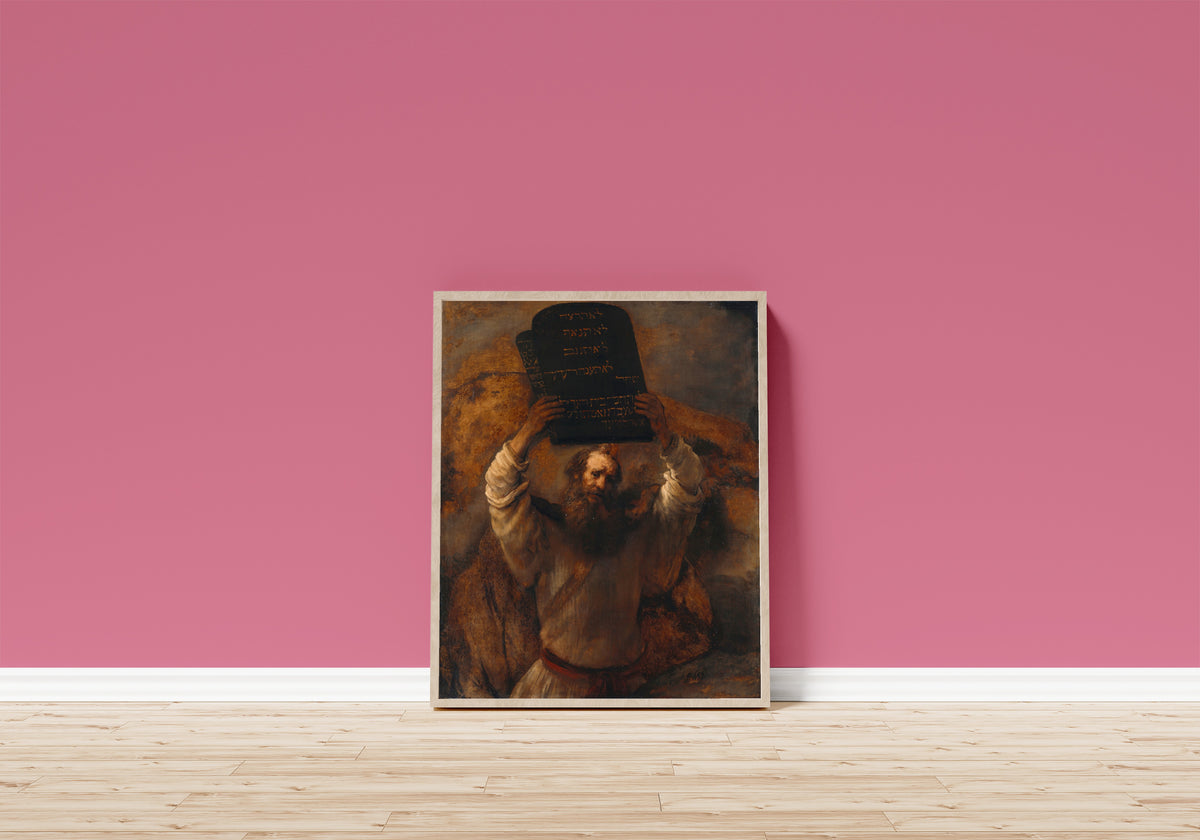 Framed art print of Moses with the Tablets of the Law by Rembrandt van Rijn, in a light wood frame leaning on a pink wall above a wooden floor