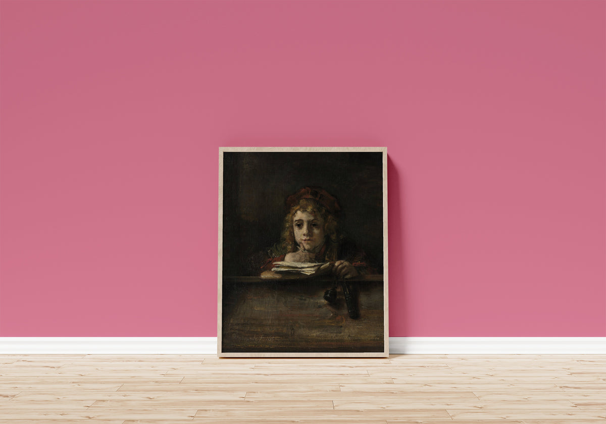 Framed art print of Titus at His Desk by Rembrandt van Rijn, in a light wood frame leaning on a pink wall above a wooden floor