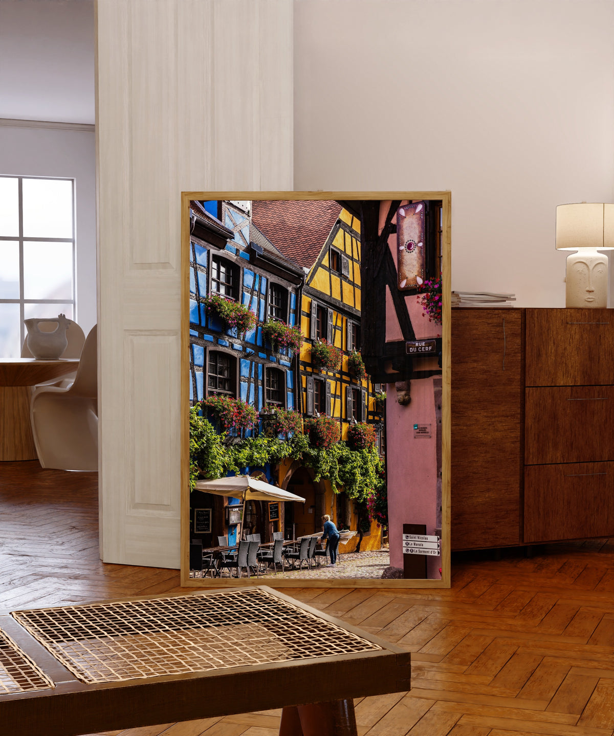 Framed art print of Riquewihr (Alsace) by Daniela Faust, in a light wood frame leaning in a warm room with a parquet floor