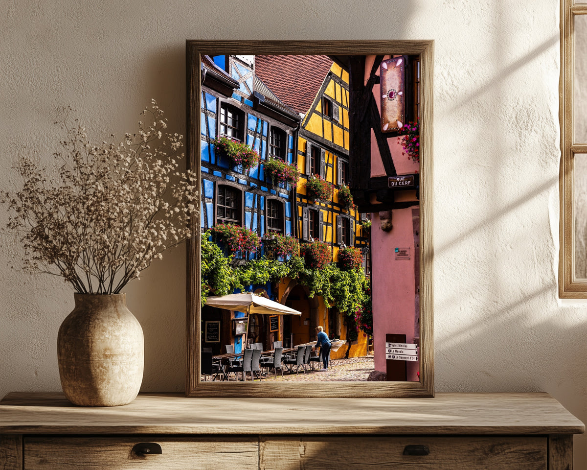 Framed art print of Riquewihr (Alsace) by Daniela Faust, in a dark wood frame leaning on a rustic console beside dried flowers