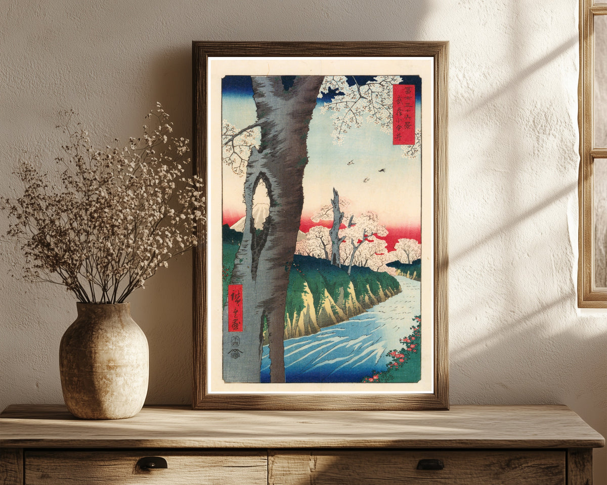 Framed art print of Koganei in Musashi Province by Utagawa Hiroshige, in a dark wood frame leaning on a rustic console beside dried flowers