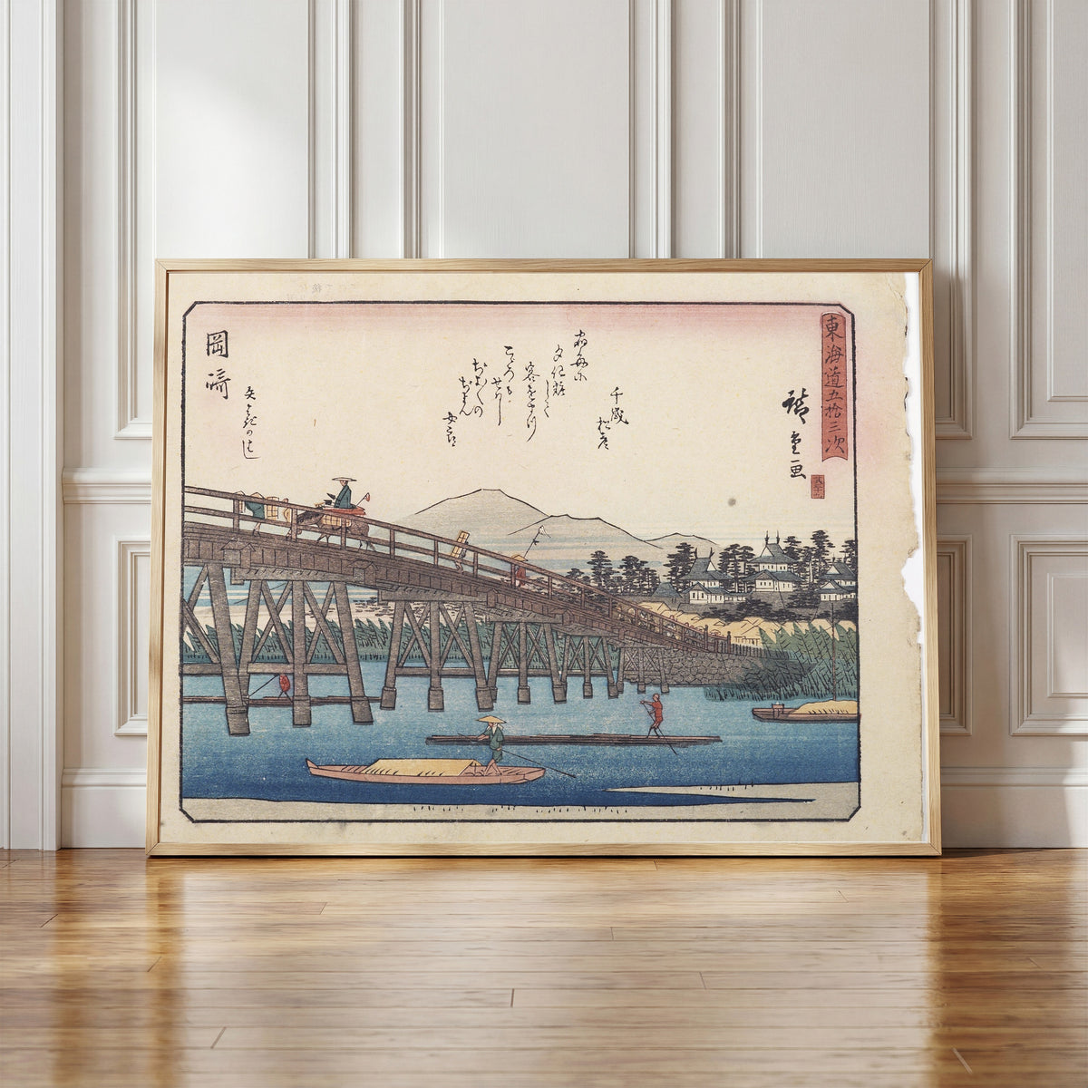 Framed art print of Kyōka Tōkaidō: Okazaki, Yahagi Bridge by Utagawa Hiroshige, in a light wood frame leaning against a panelled white wall