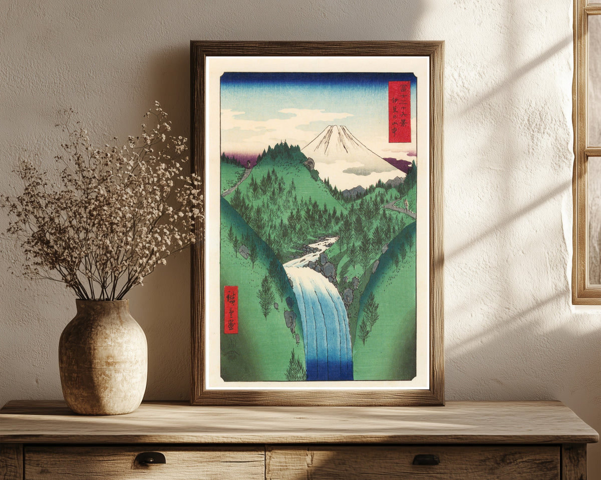 Framed art print of The Izu Mountains by Utagawa Hiroshige, in a dark wood frame leaning on a rustic console beside dried flowers
