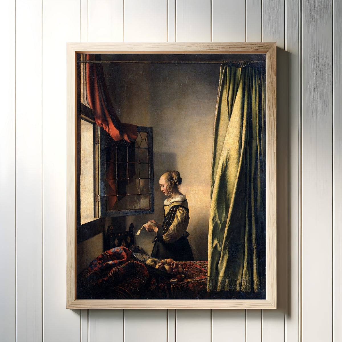 Framed art print of Young Woman Reading a Letter at an Open Window by Johannes Vermeer, in a light wood frame on a white panelled wall