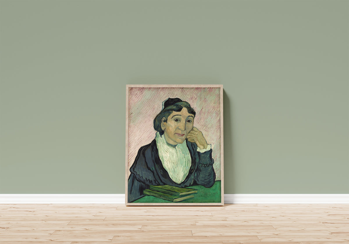 Framed art print of L'Arlésienne (Madame Ginoux) by Vincent van Gogh, in a light wood frame leaning on a sage green wall above a wooden floor