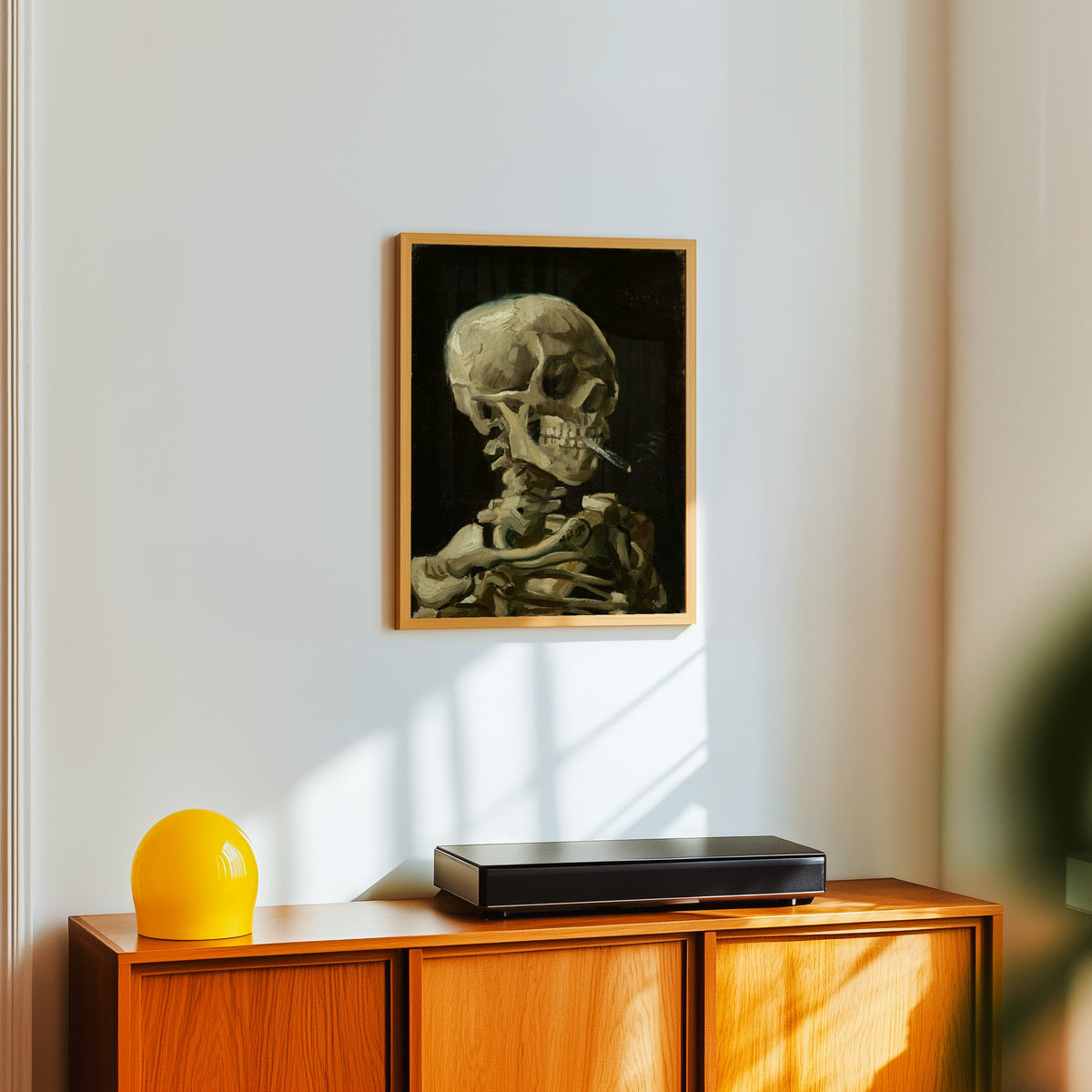 Framed art print of Skull with Burning Cigarette by Vincent van Gogh, in a light wood frame on a white wall above an orange sideboard
