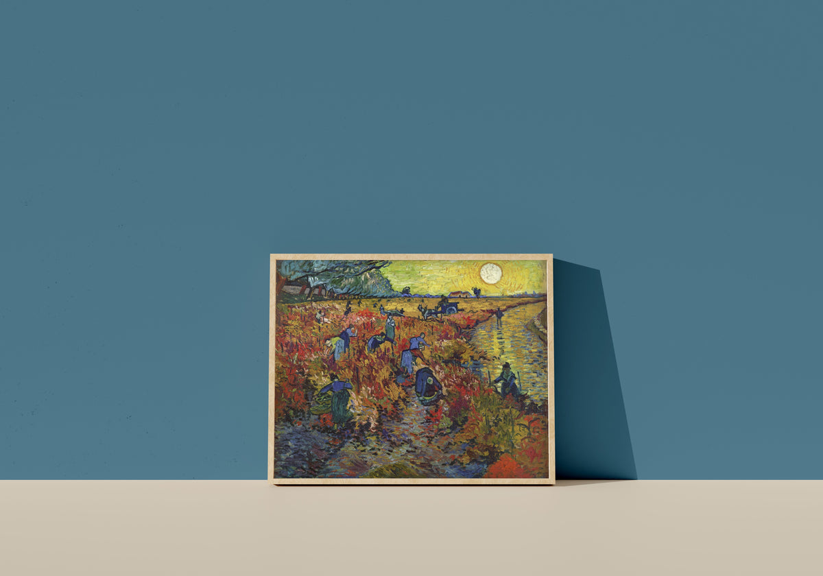 Framed art print of The Red Vineyard by Vincent van Gogh, in a light wood frame leaning on a blue wall