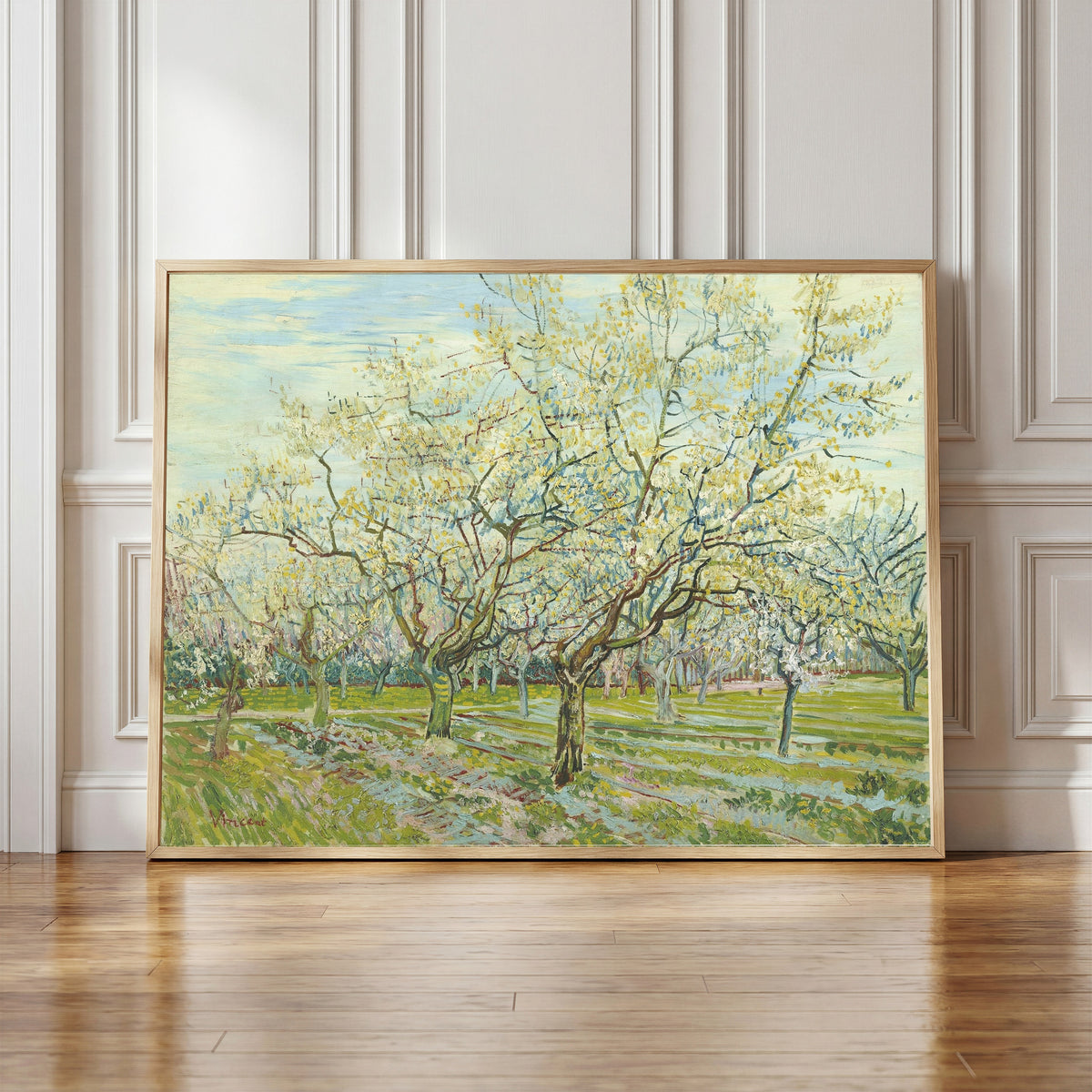 Framed art print of The White Orchard by Vincent van Gogh, in a light wood frame leaning against a panelled white wall