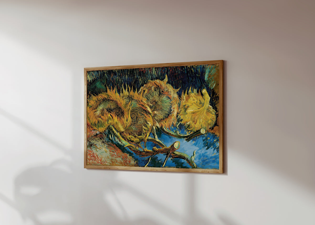 Framed art print of Sunflowers by Vincent van Gogh, in a light wood frame on a sunlit white wall, seen at a slight angle