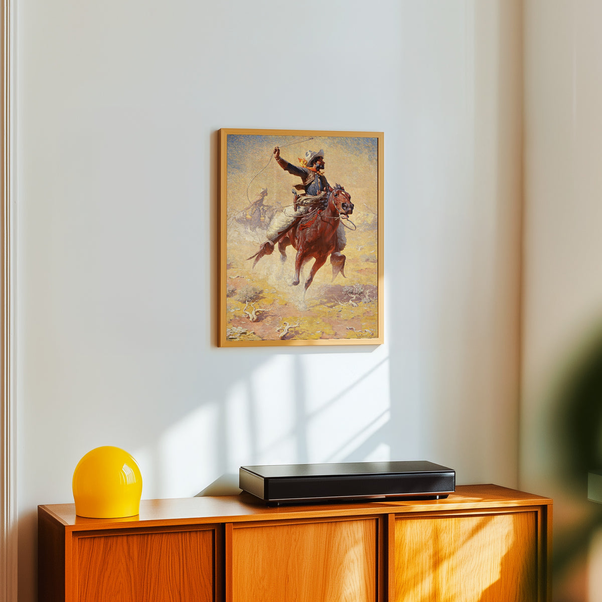 Framed art print of The Roping by William Robinson Leigh, in a light wood frame on a white wall above an orange sideboard