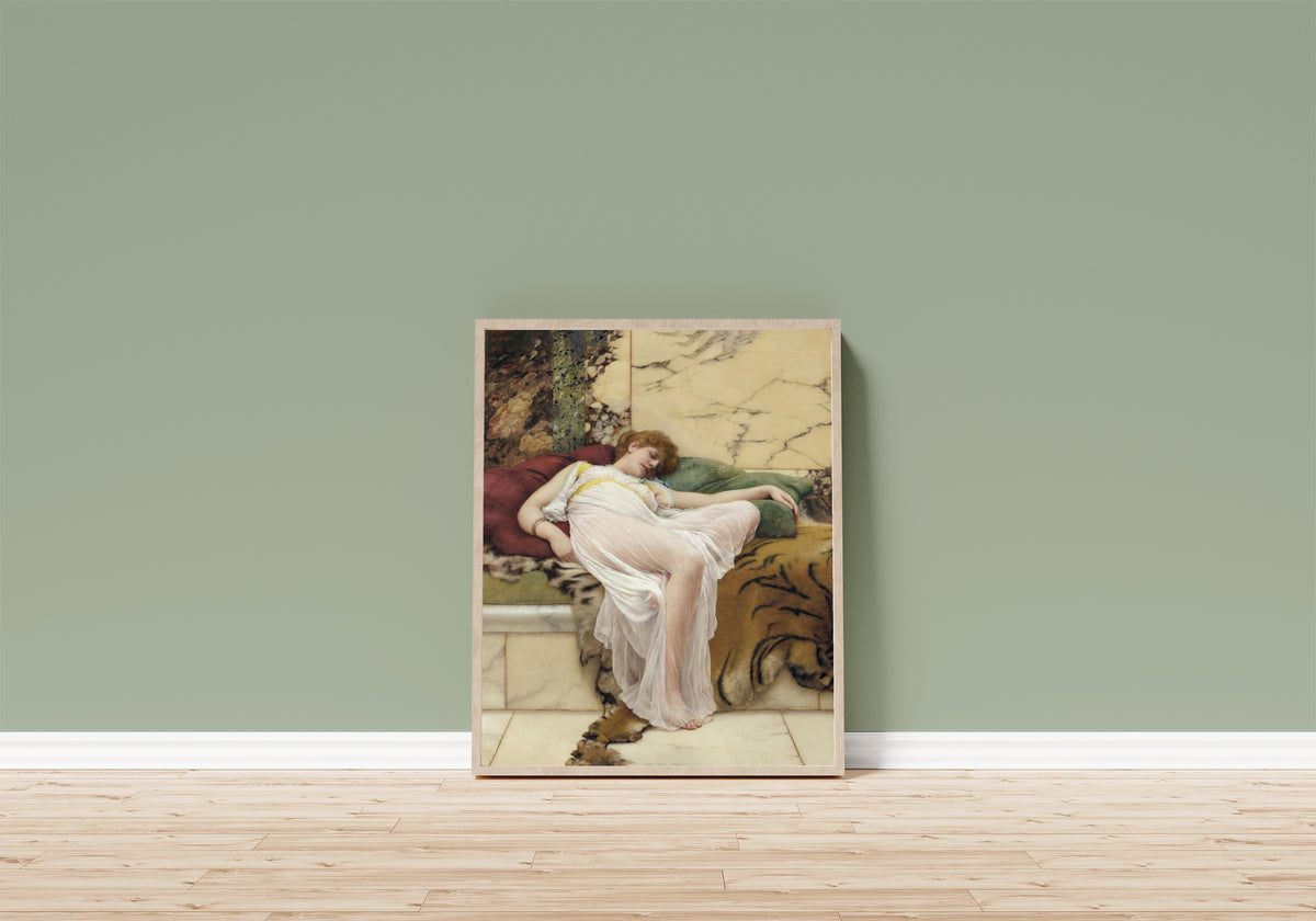 Framed art print of A Siesta by John William Godward, in a light wood frame leaning on a sage green wall above a wooden floor