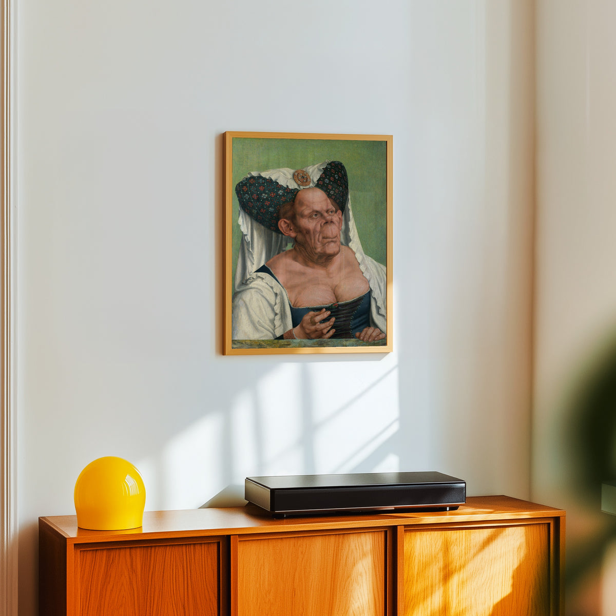 Framed art print of An Old Woman (The Ugly Duchess) by Quinten Massys, in a light wood frame on a white wall above an orange sideboard
