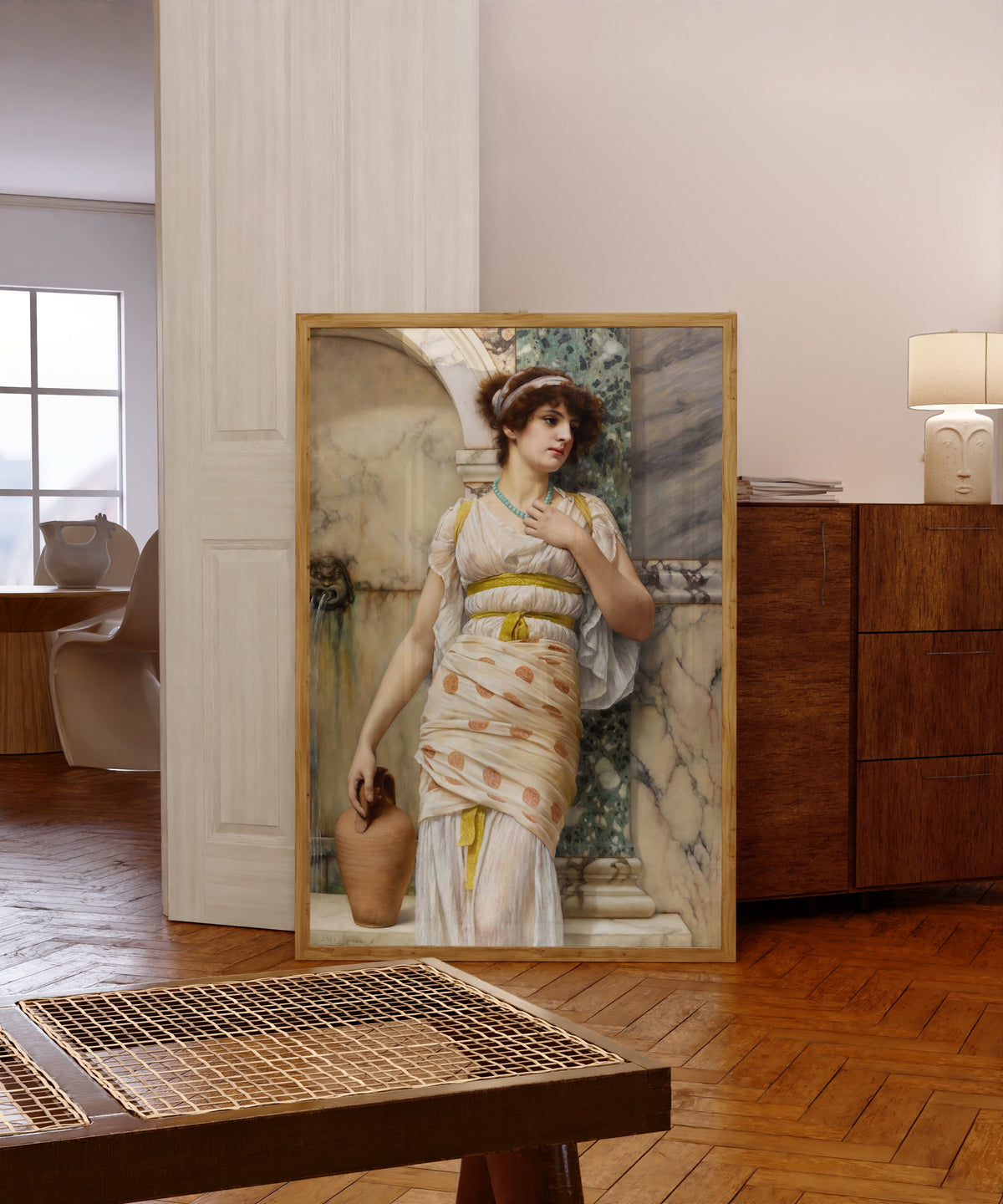 Framed art print of At the Fountain by John William Godward, in a light wood frame leaning in a warm room with a parquet floor