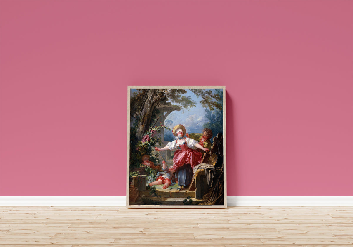 Framed art print of Blind-Man’s Buff by Jean-Honoré Fragonard, in a light wood frame leaning on a pink wall above a wooden floor