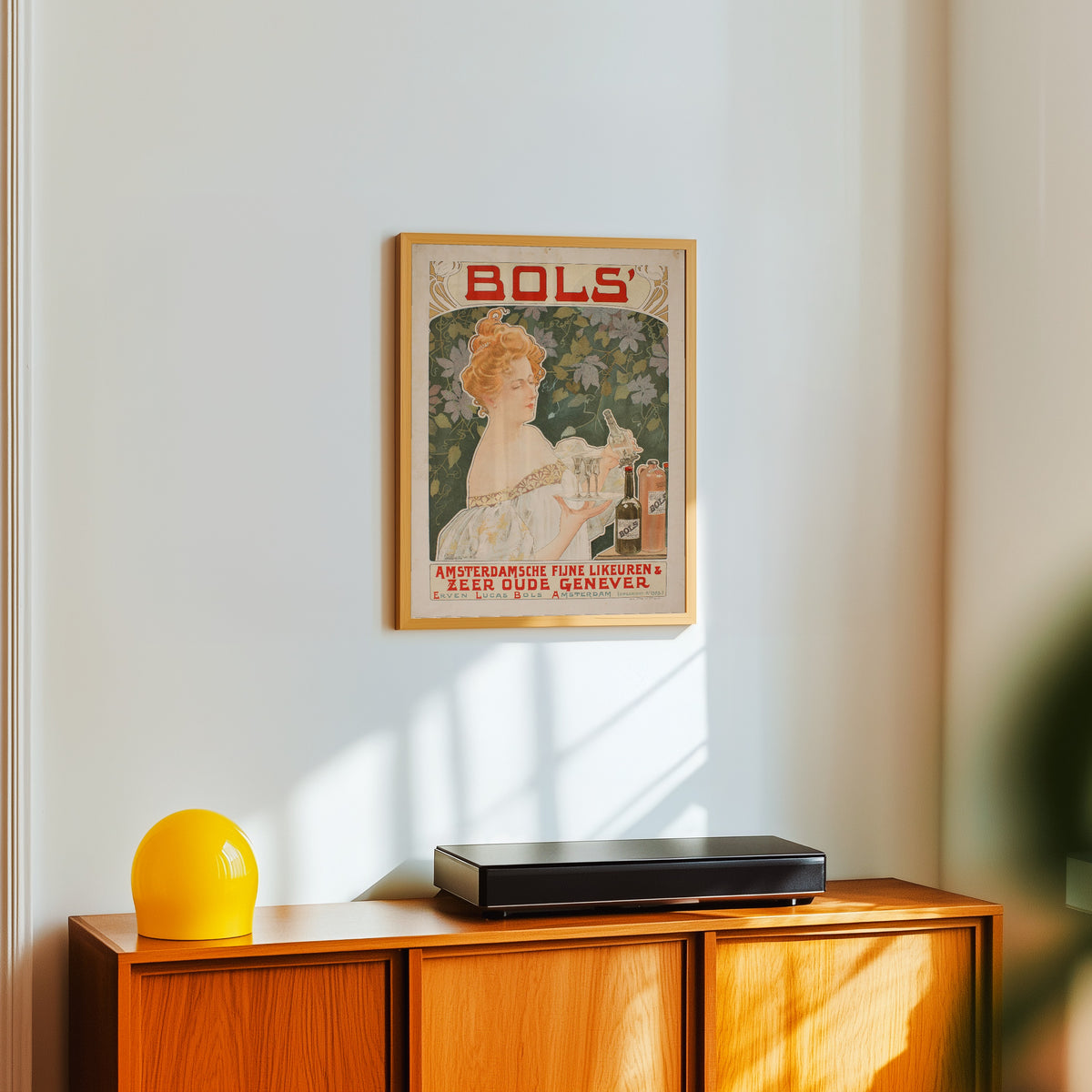 Framed art print of Bols' Amsterdamsche Fijne Likeuren by Henri Privat-Livemont, in a light wood frame on a white wall above an orange sideboard