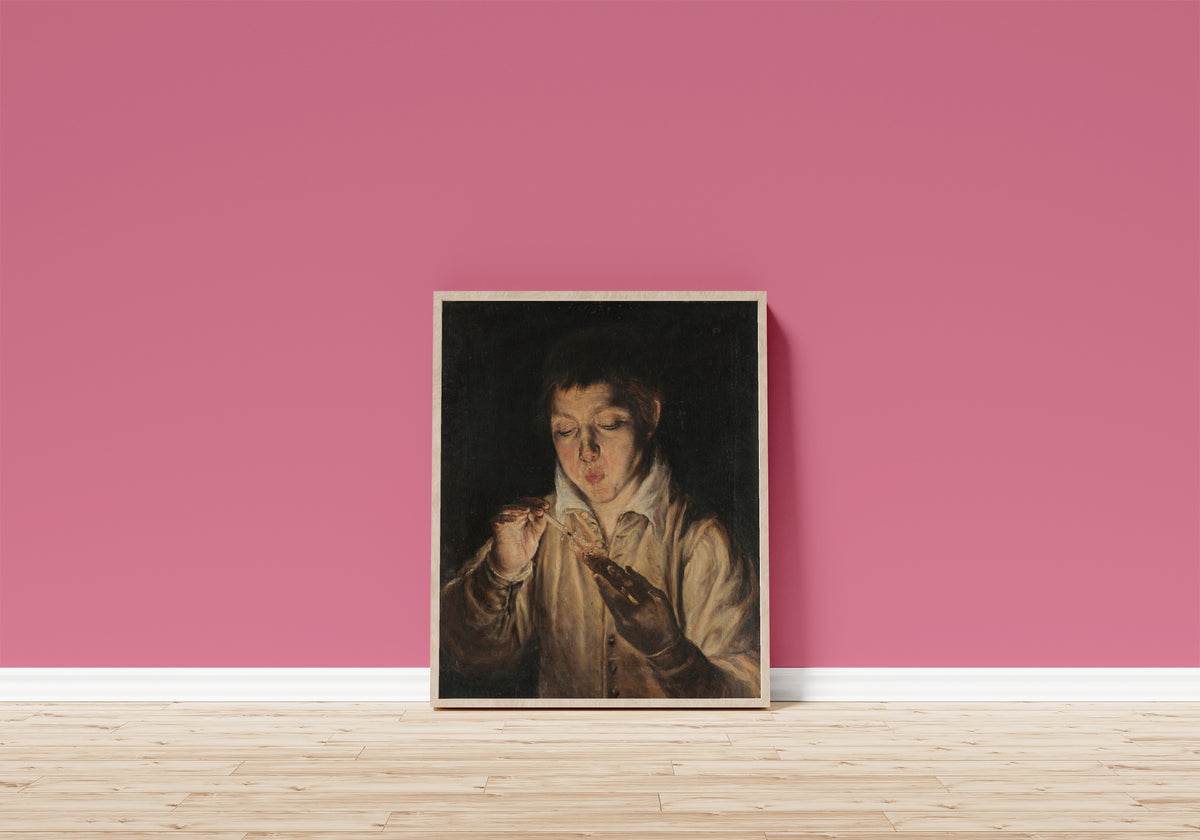 Framed art print of Boy Blowing an Ember by El Greco, in a light wood frame leaning on a pink wall above a wooden floor
