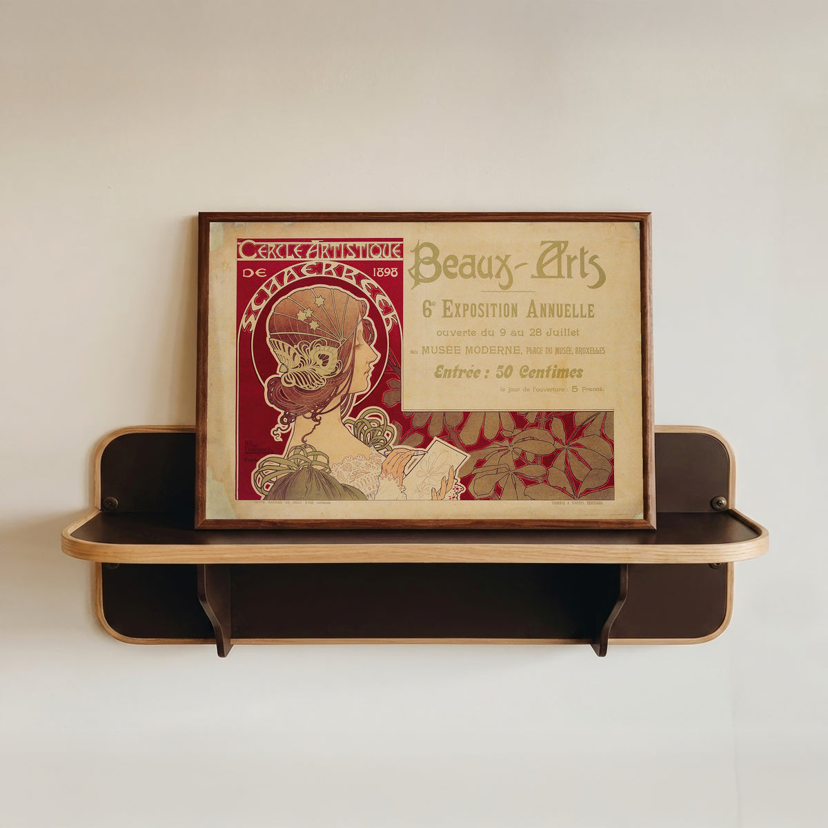 Framed art print of Cercle Artistique de Schaerbeek (1898) by Henri Privat-Livemont, in a dark wood frame resting on a wall shelf