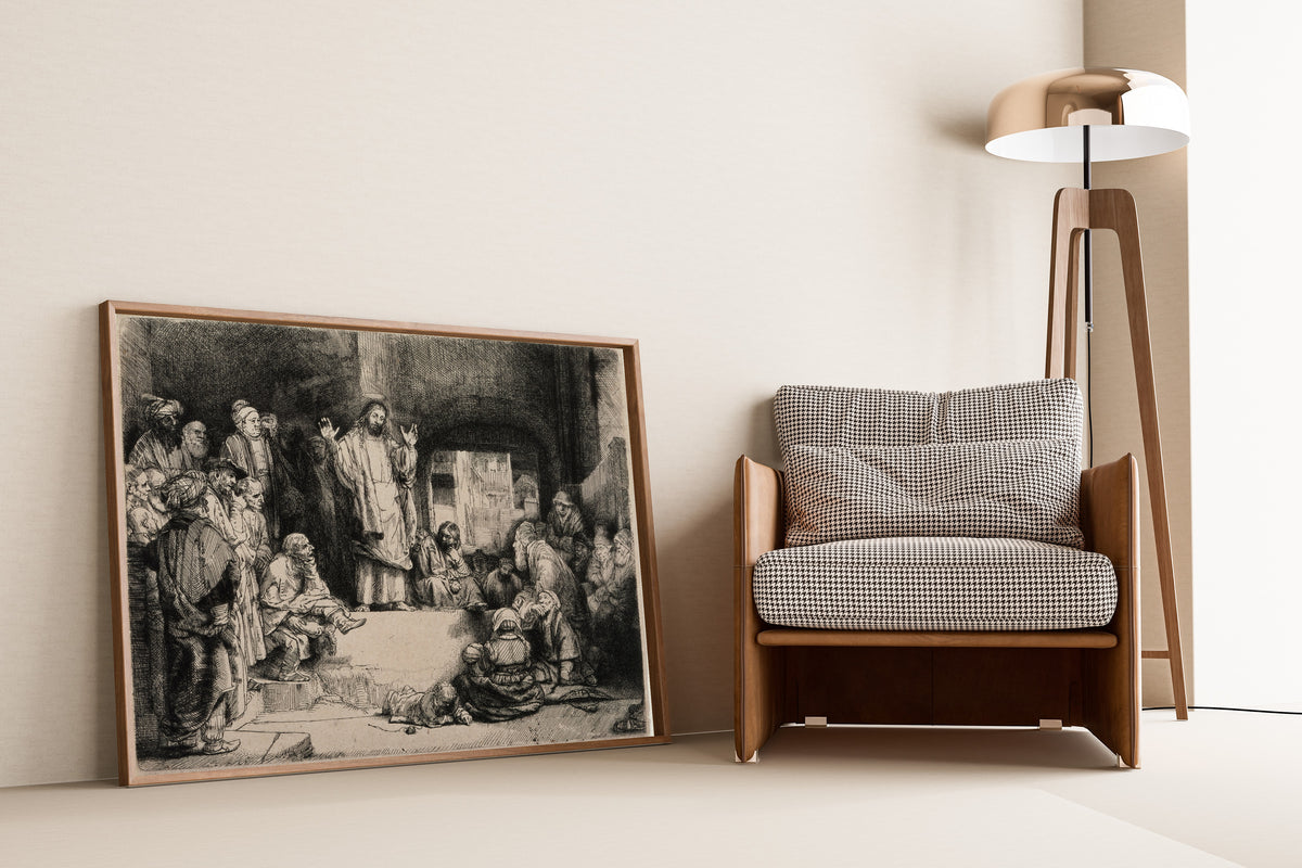 Framed art print of Christ Preaching, called La Petite Tombe by Rembrandt van Rijn, leaning against a wall beside an armchair and a floor lamp