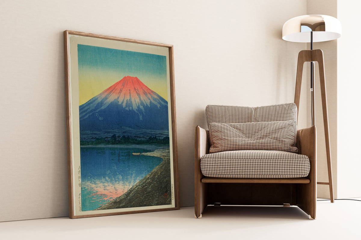 Framed art print of Daybreak over Lake Yamanaka by Kawase Hasui, leaning against a wall beside an armchair and a floor lamp