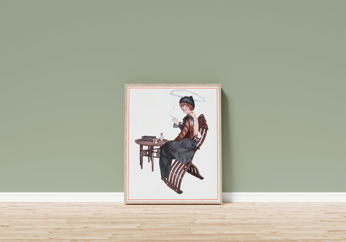 Framed art print of De la Brune à la Blonde, Pl. 4: L'Auréole de… by Raphael Kirchner, in a light wood frame leaning on a sage green wall above a wooden floor
