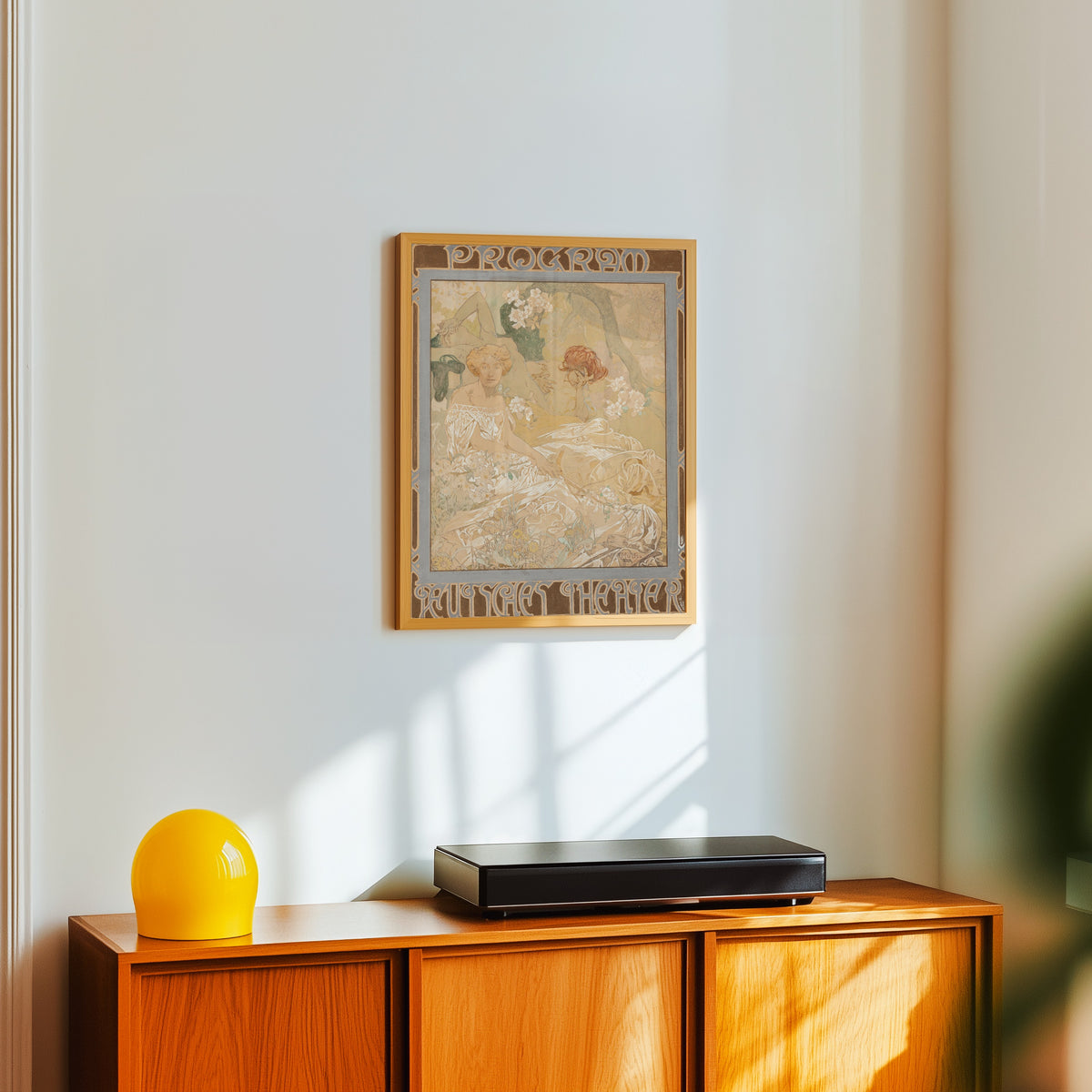 Framed art print of Deutsches Schauspiel-Haus, Theater Program by Alphonse Mucha, in a light wood frame on a white wall above an orange sideboard