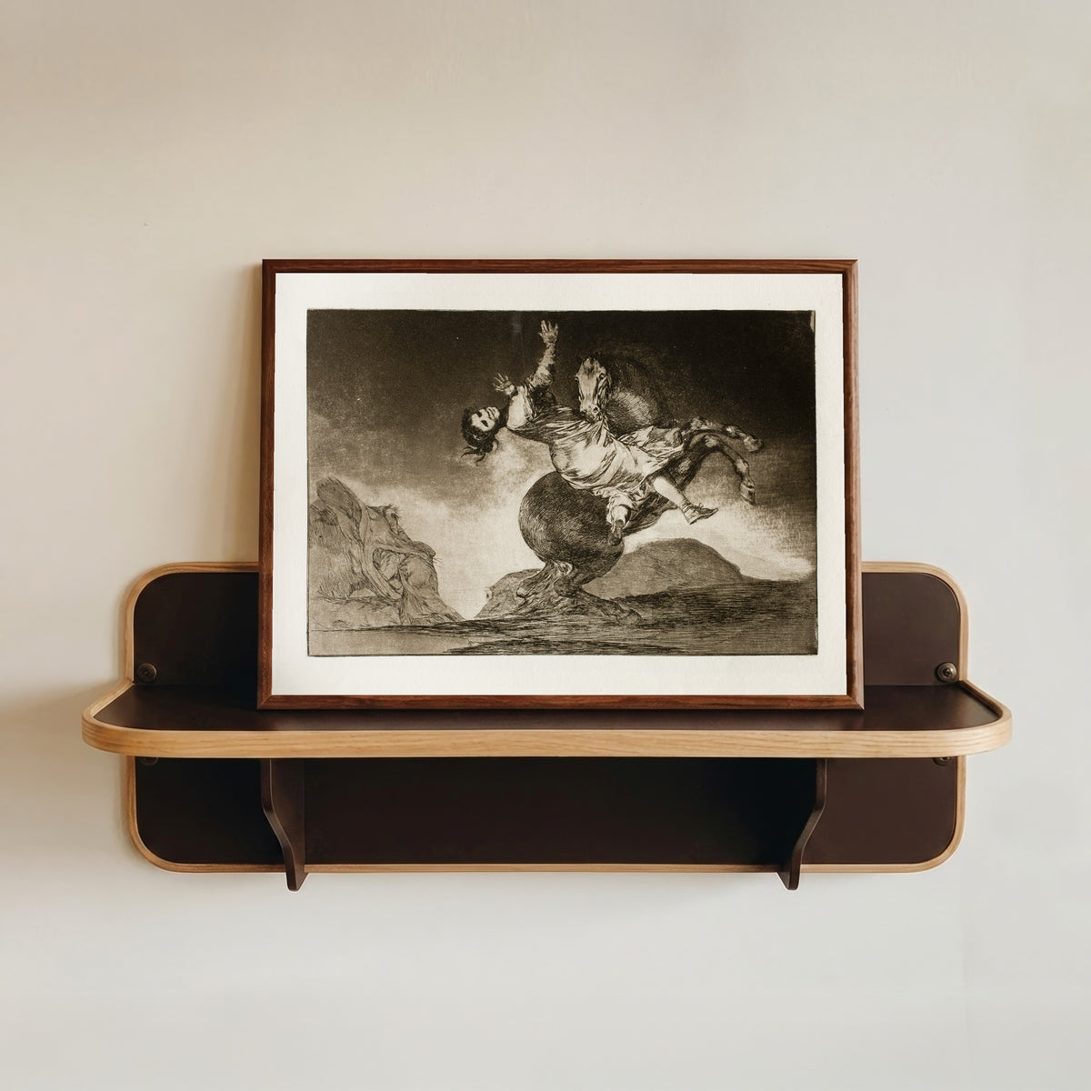 Framed art print of El caballo raptor (The Horse Abductor), from Los Disparates by Francisco de Goya, in a dark wood frame resting on a wall shelf