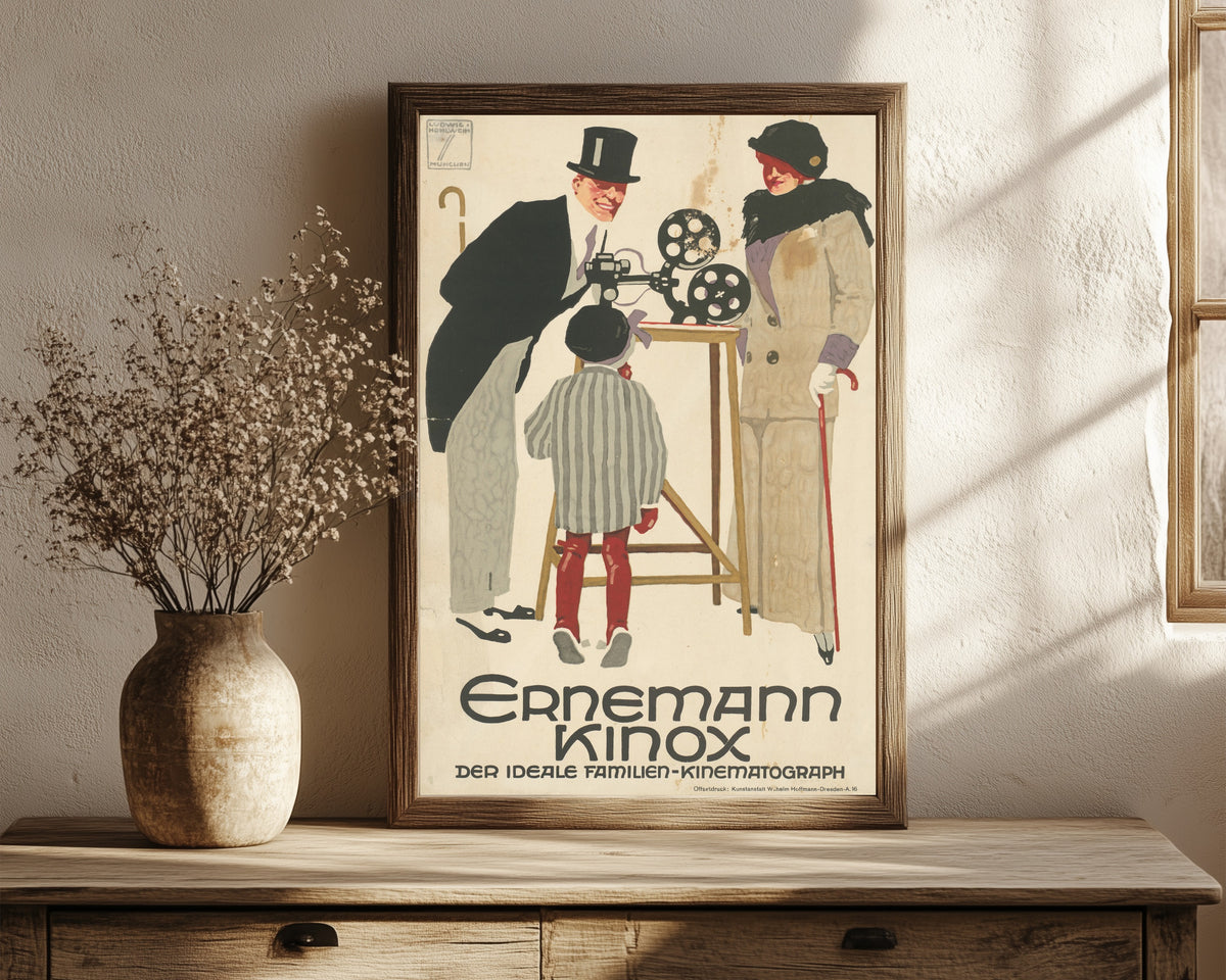 Framed art print of Ernemann Kinox: Der ideale Familien-Kinematograph by Ludwig Hohlwein, in a dark wood frame leaning on a rustic console beside dried flowers