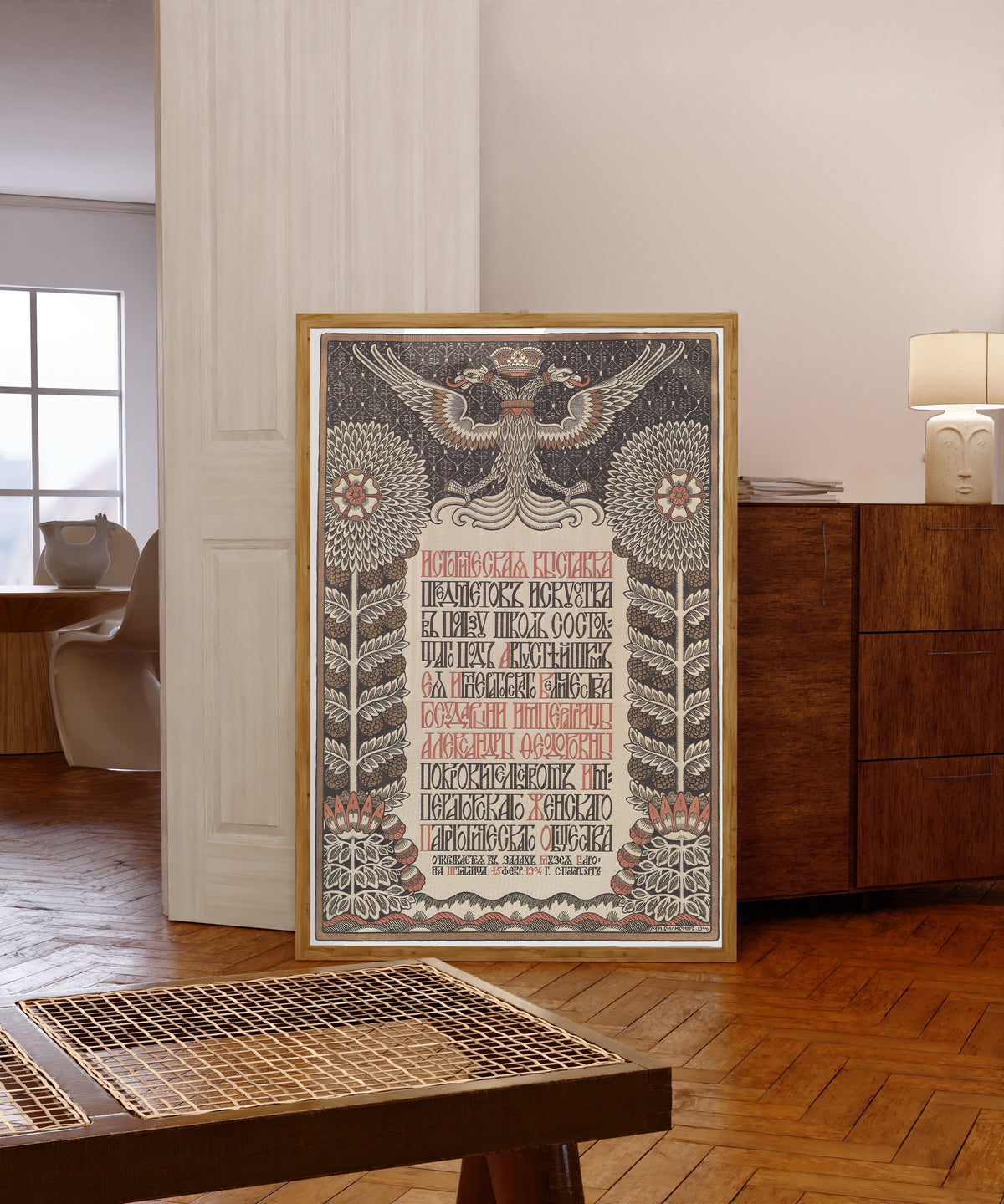 Framed art print of Historical Exhibition of Art Objects in Aid of Schools by Ivan Bilibin, in a light wood frame leaning in a warm room with a parquet floor
