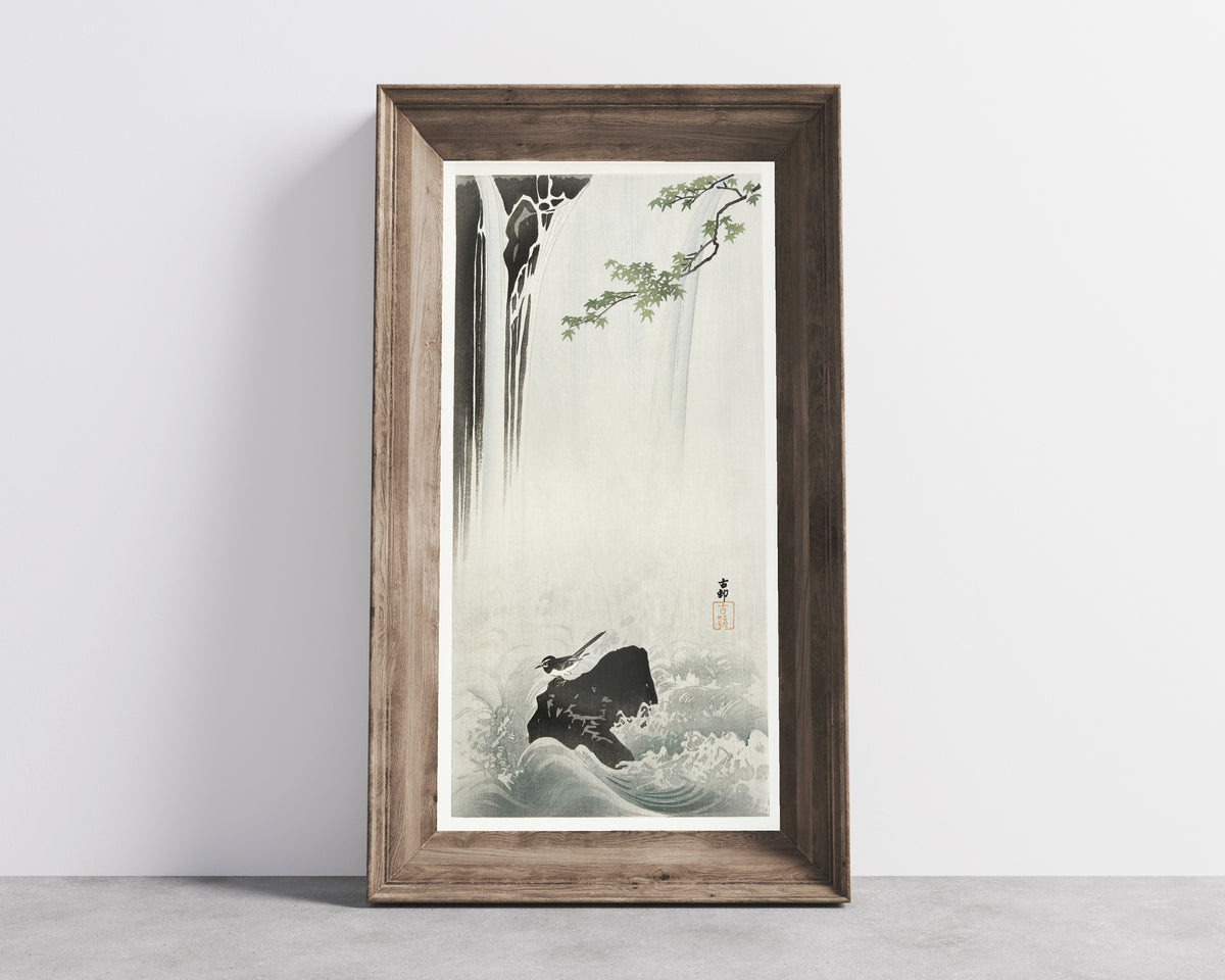 Framed art print of Japanese Wagtail at a Waterfall by Ohara Koson, in a wide wooden frame leaning against a concrete wall