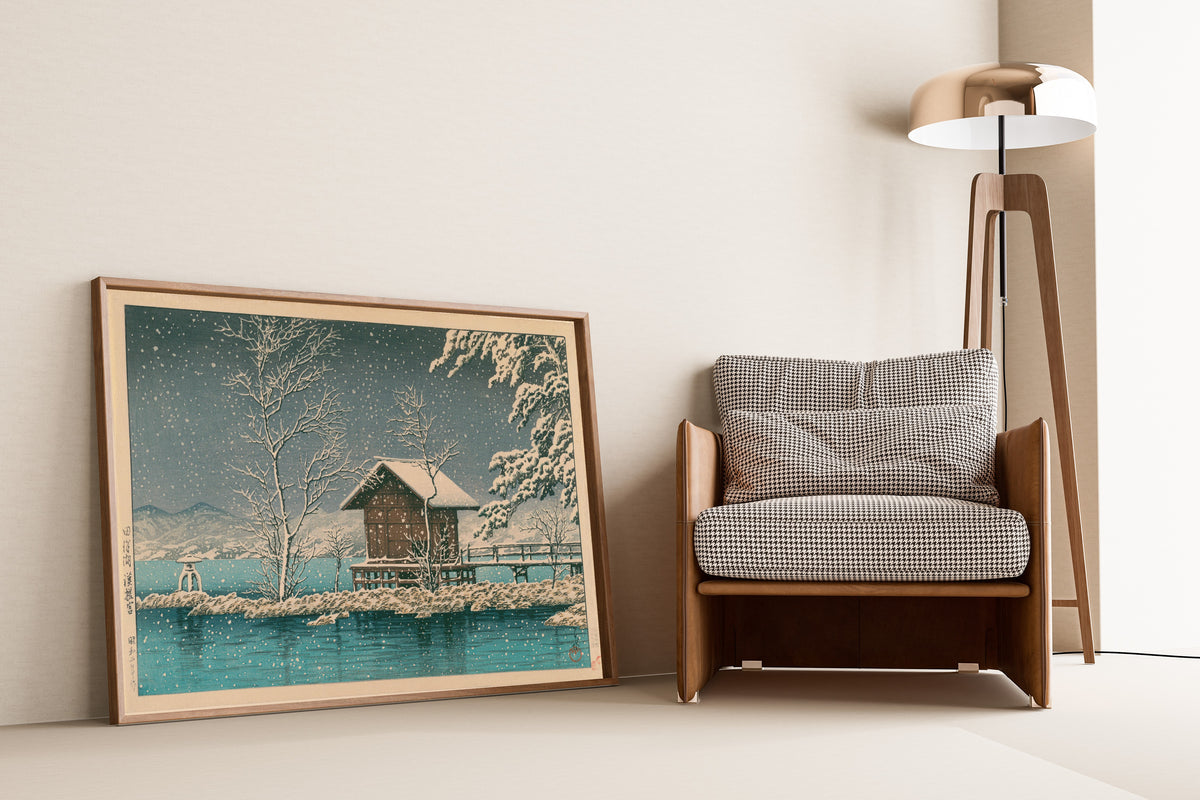 Framed art print of Kansanomiya Shrine at Lake Tazawa by Kawase Hasui, leaning against a wall beside an armchair and a floor lamp