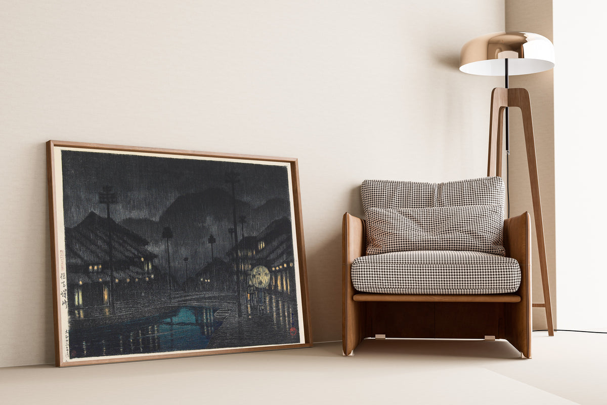 Framed art print of Kinosaki in Tajima Province by Kawase Hasui, leaning against a wall beside an armchair and a floor lamp