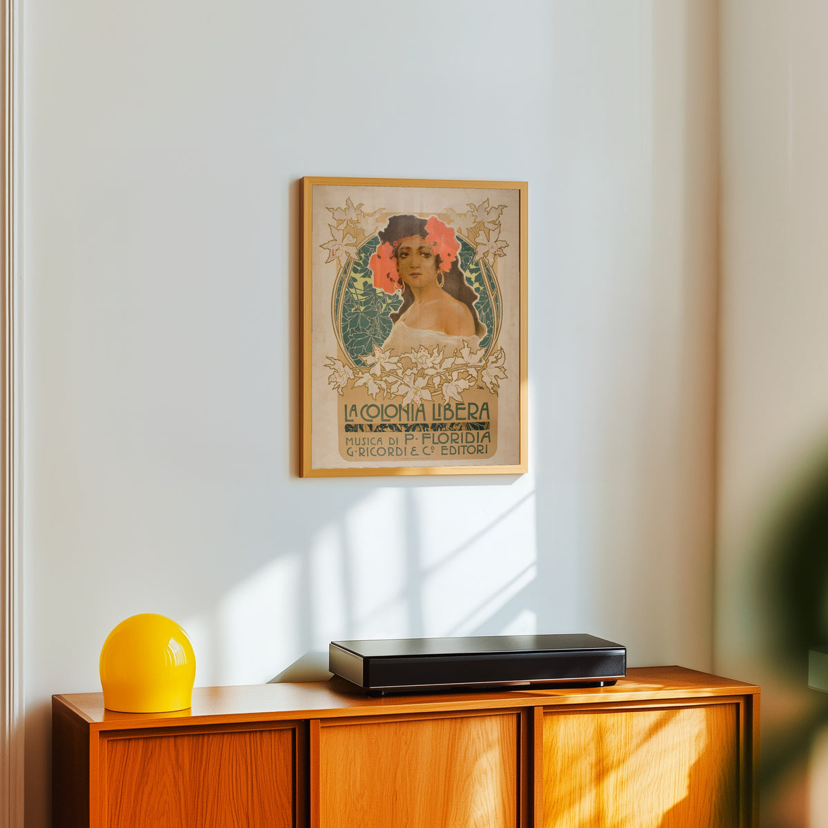 Framed art print of La Colonia Libera by Leopoldo Metlicovitz, in a light wood frame on a white wall above an orange sideboard
