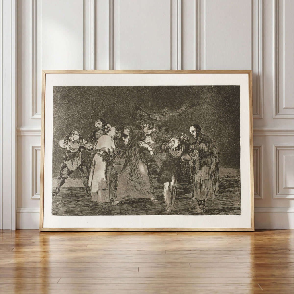 Framed art print of Las exhortaciones (The Exhortations), from Los Disparates by Francisco de Goya, in a light wood frame leaning against a panelled white wall