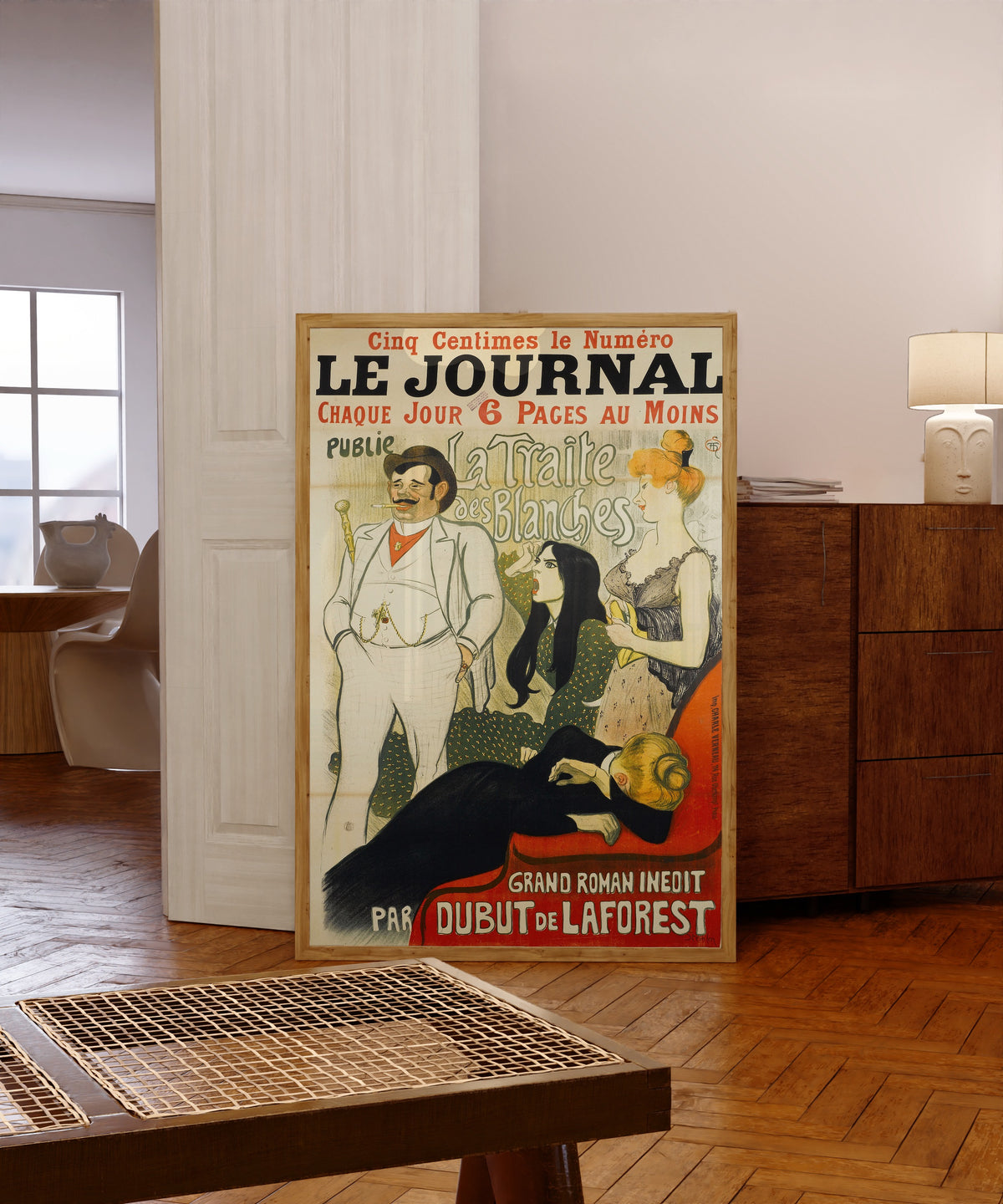 Framed art print of Le Journal: La Traite des Blanches by Théophile Steinlen, in a light wood frame leaning in a warm room with a parquet floor