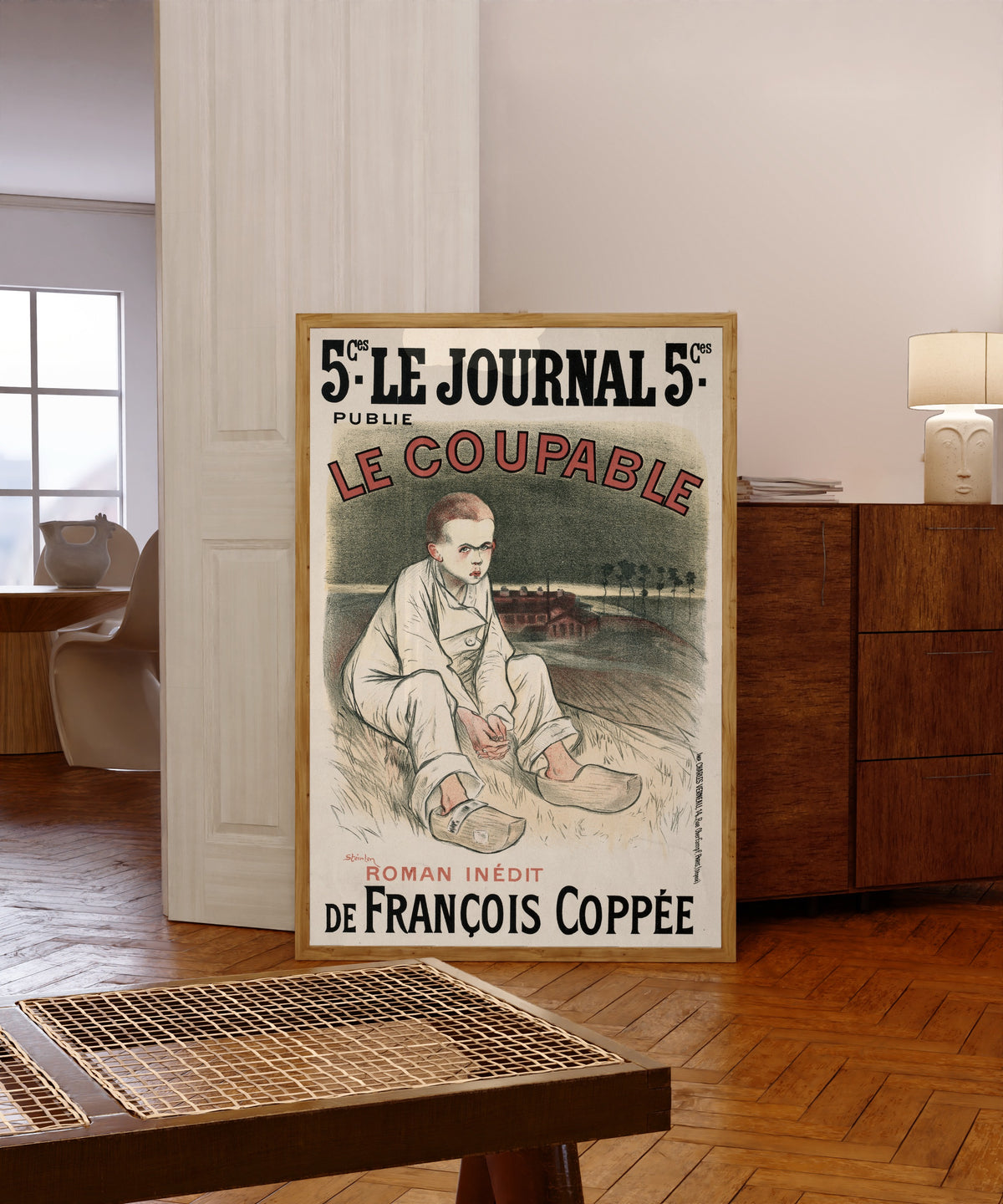 Framed art print of Le Journal: Le Coupable by Théophile Steinlen, in a light wood frame leaning in a warm room with a parquet floor