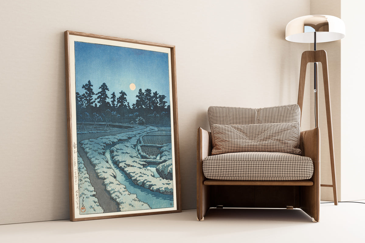 Framed art print of Moon at Ayashi, Miyagi Prefecture by Kawase Hasui, leaning against a wall beside an armchair and a floor lamp