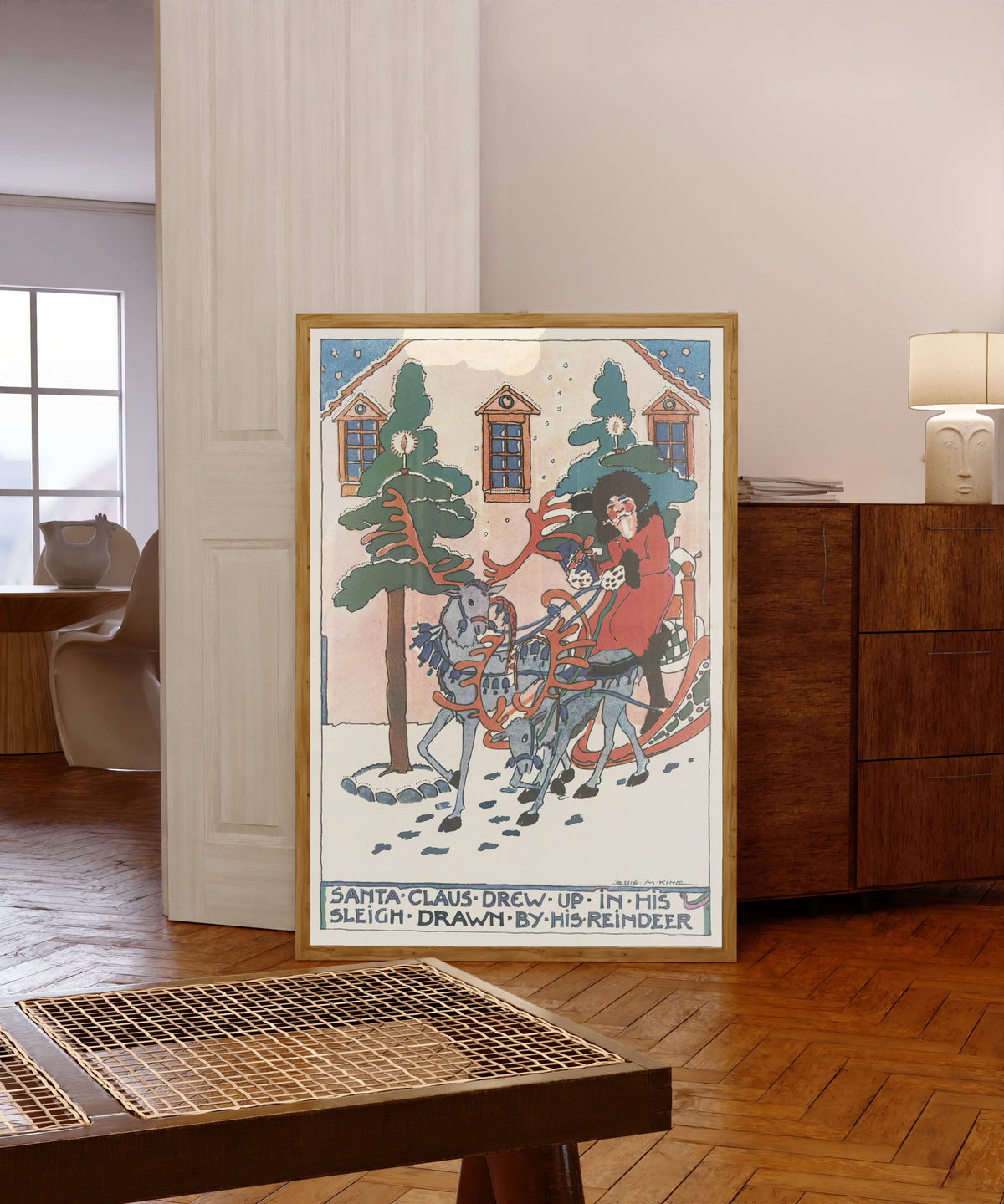 Framed art print of Mummy's Bedtime Story Book: Santa Claus Drew Up in… by Jessie M. King, in a light wood frame leaning in a warm room with a parquet floor