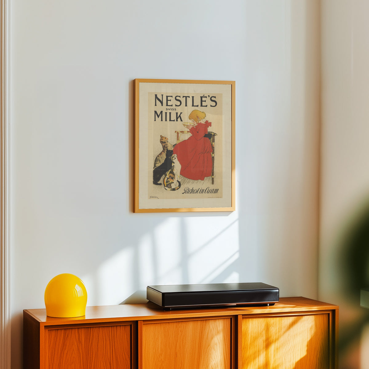 Framed art print of Nestlé’s Swiss Milk, Richest in Cream by Théophile Steinlen, in a light wood frame on a white wall above an orange sideboard