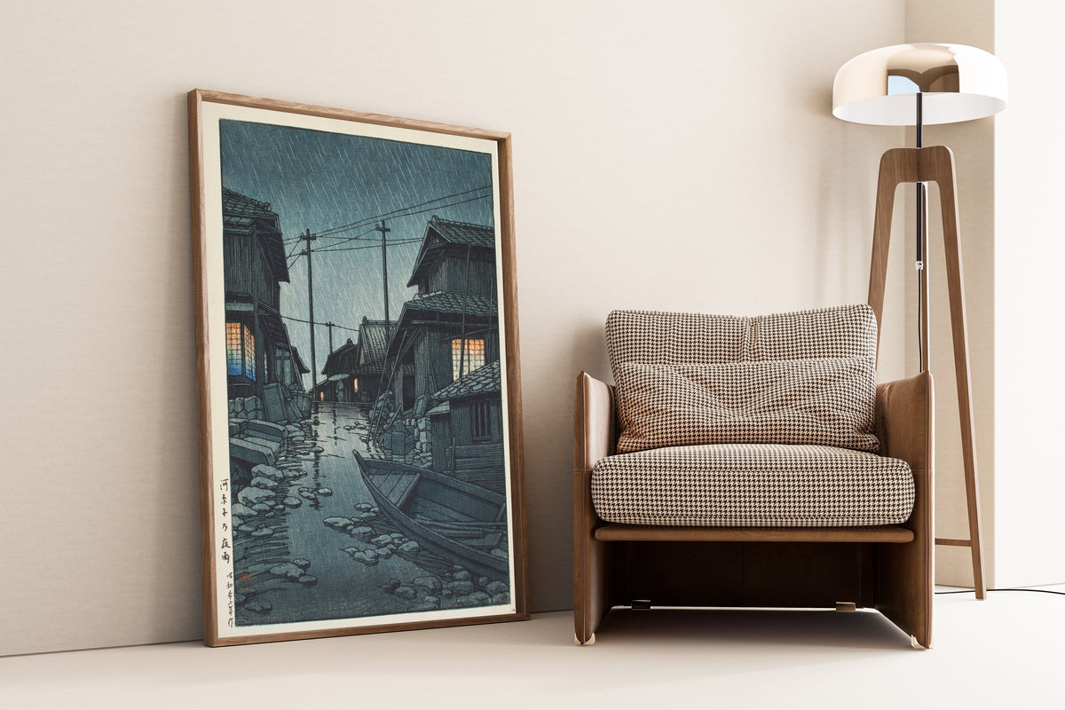 Framed art print of Night Rain at Kawarago by Kawase Hasui, leaning against a wall beside an armchair and a floor lamp